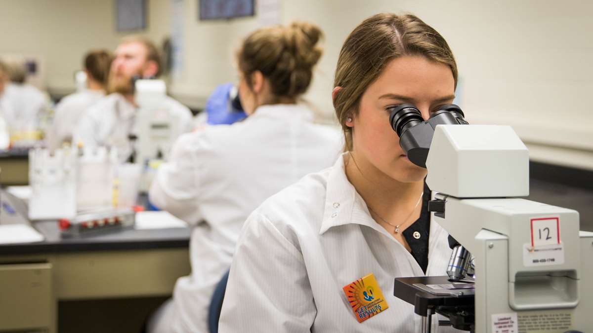 A young female scientist in a white lab coat looking through a microscope.