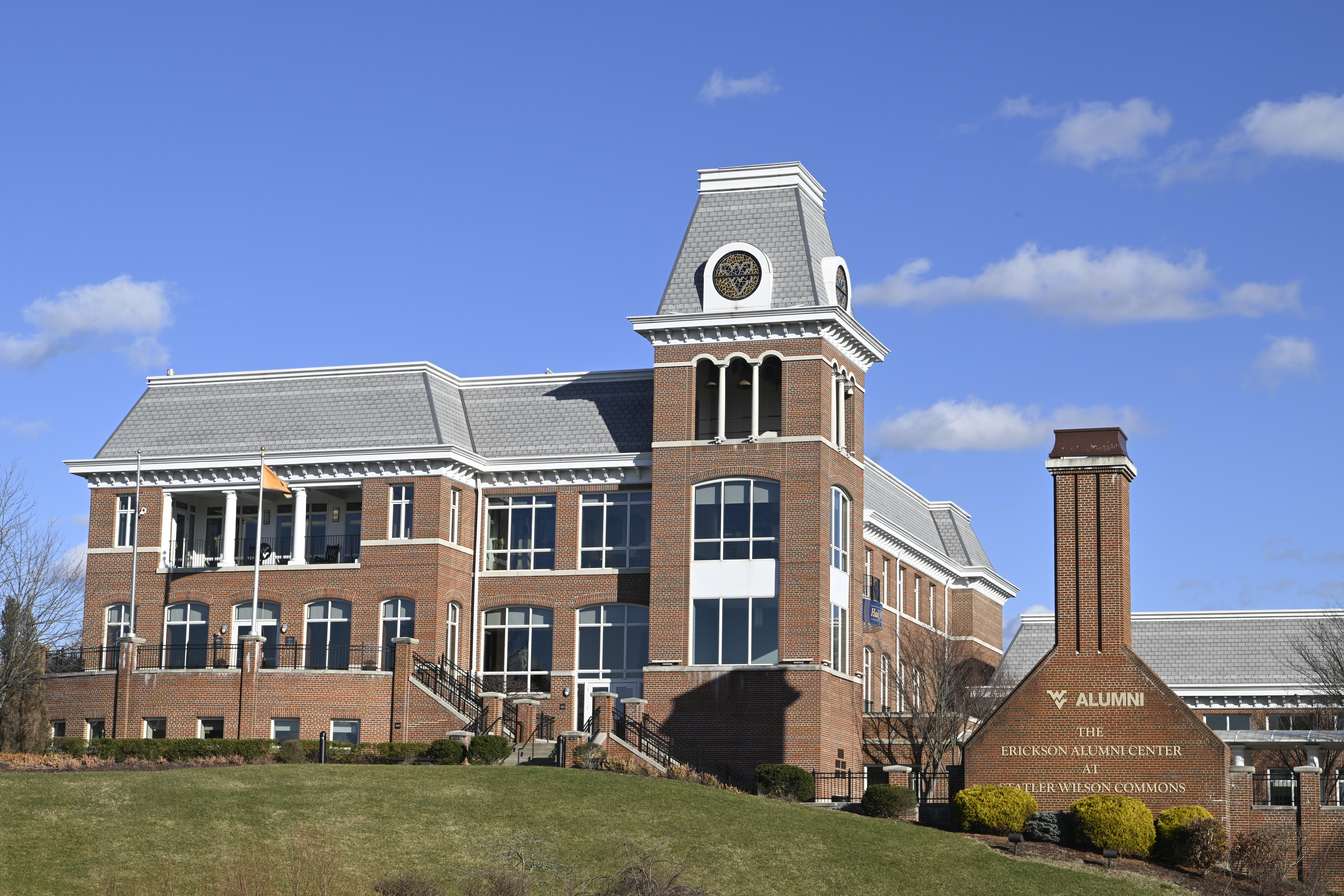 A wide shot of the Erickson Alumni Center is seen against a mostly clear blue sky. The red brick building has a wing to the left; a tower near center capped by a “Flying WV” circular stained glass window near its top; and a brick wall with a gold WVU Alumni wordmark and text beneath it that reads, “The Erickson Alumni Center at Statler Wilson Commons” in gold lettering.
