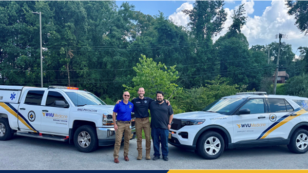  A group of three smiling individuals stand in front of a white emergency SUV with a light bar on the top and the "WVU Medicine, Emergency Rescue" logo on the side.