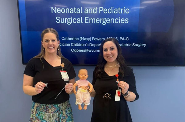 Two Physician Assistant female preceptors holding a baby doll in front of a screen that reads, "Neonatal and Pediatric Surgical Emergencies"